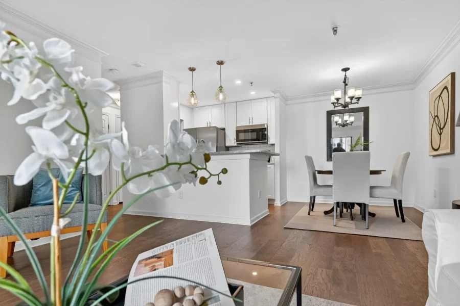 Bright open-plan living area with a white kitchen, island, and a dining table with four upholstered chairs; white orchids in the foreground.