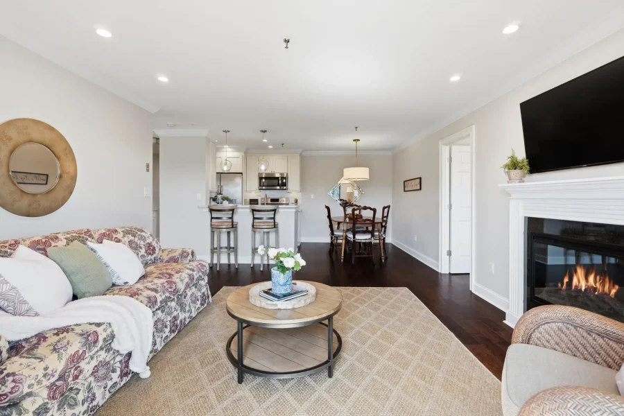 Open-concept living room with a floral-patterned sofa, round coffee table, beige walls, and a fireplace; kitchen and dining area visible in back.