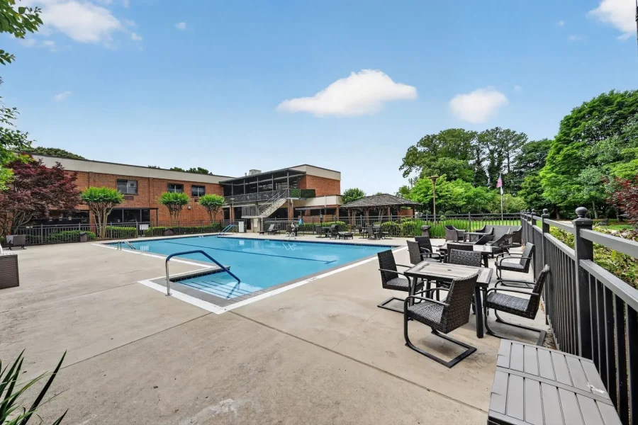 Outdoor hotel-style swimming pool with a bright blue water, concrete deck, and a metal fence; lounge chairs and tables line the right side, brick building in the background, and green trees under a clear sky.