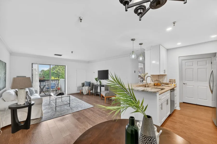 Open-concept living room with a white kitchen, hardwood floors, and a balcony view through sliding doors; seating area includes sofa, chairs, and a coffee table on a gray rug.