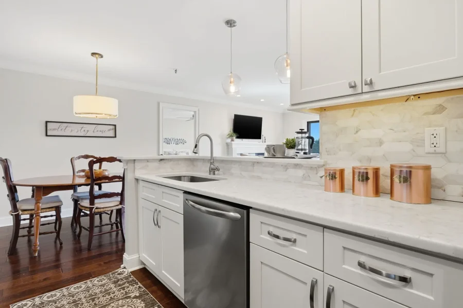 Bright modern kitchen with white shaker cabinets, marble countertops, and copper canisters on the island; a dining area with wooden chairs is visible in the background.