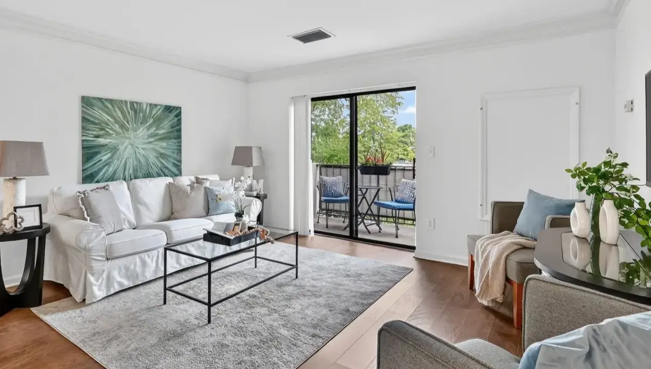 Bright living room with white walls, a white sofa and gray pillows, glass coffee table, and a balcony door with outdoor seating visible outside.