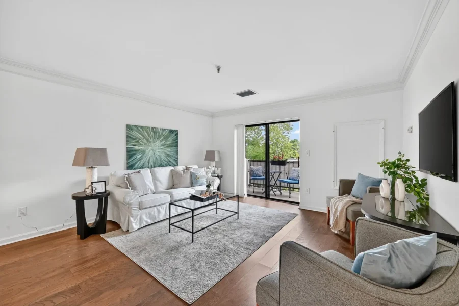Bright living room with white walls, a white sofa and gray pillows, glass coffee table, and a balcony door with outdoor seating visible outside.