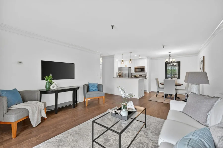 Open-concept living room with gray sofas, a wall-mounted TV, and a glass coffee table; kitchen and dining area in back.
