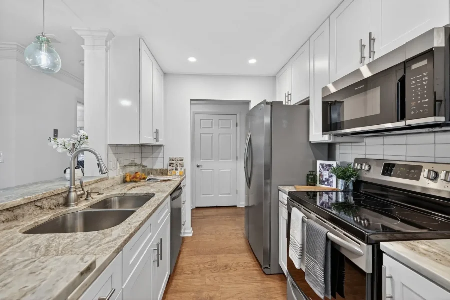 Modern white kitchen with marble countertops, stainless steel appliances (refrigerator, microwave, oven), and a double sink along a long island, seen in a narrow galley layout toward a back doorway.