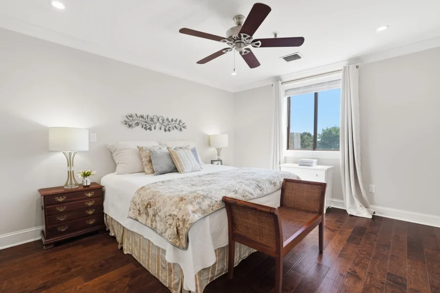 Bright bedroom with a white bed, floral quilt, two matching nightstands with lamps, and a ceiling fan. A large window with white curtains lets in daylight.