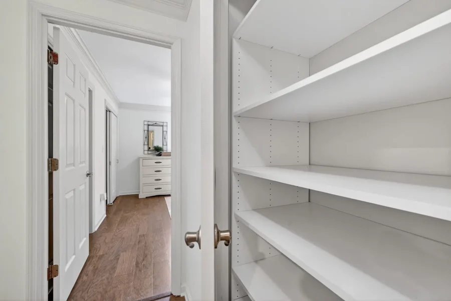 Narrow hallway with white paneled doors; open door reveals white built-in shelving unit on the right and a hardwood floor leading to a dresser with a mirror at the far end.