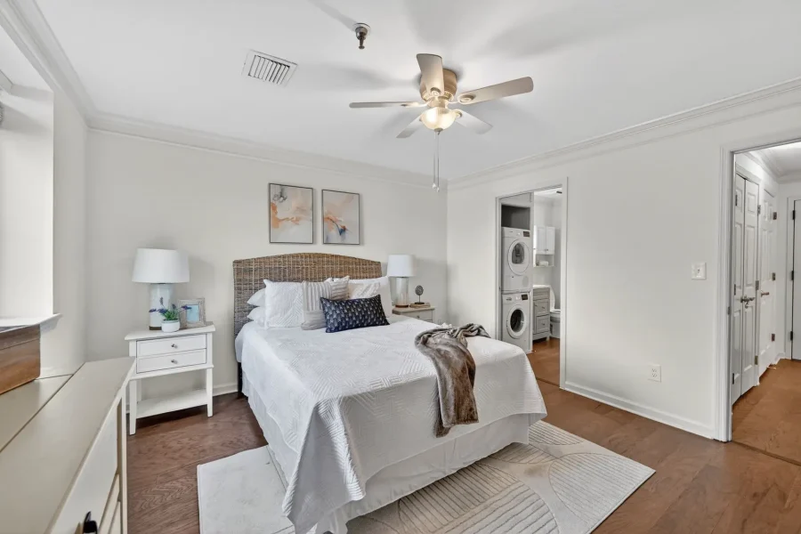 Bright bedroom with white bedding, wicker headboard, and matching nightstands; laundry area visible through open doorway in back.