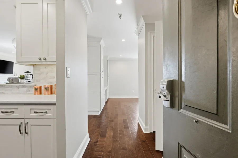 Open kitchen with white shaker cabinets on left, marble backsplash, and dark wood floor; stainless steel fridge on the right, hallway ahead.
