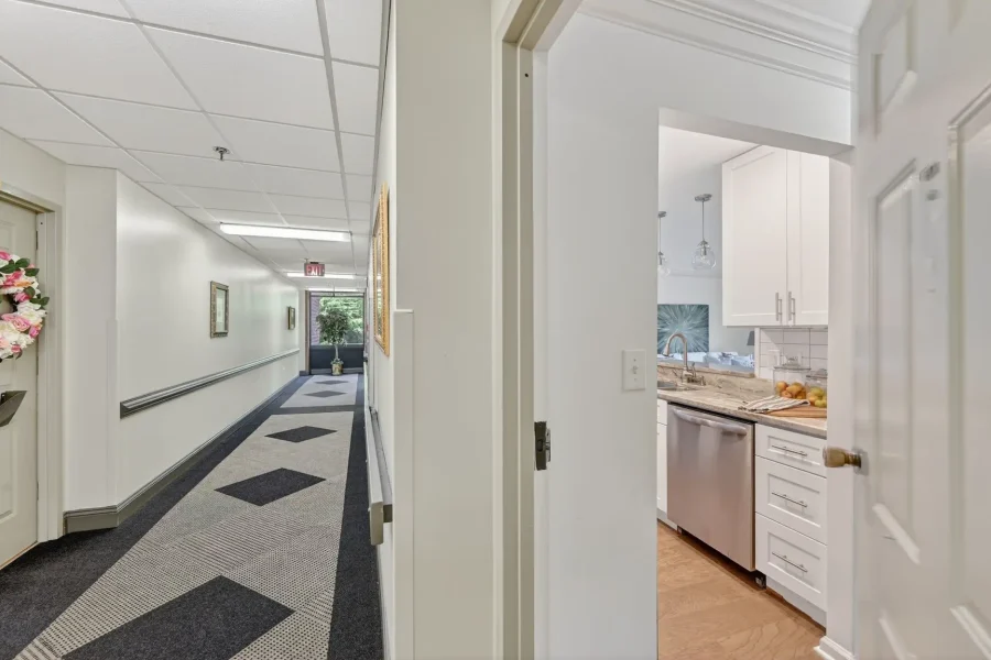 Long, well-lit hallway with white walls, a patterned carpet, and a kitchen doorway on the right showing white cabinets and a stainless dishwasher at the counter.