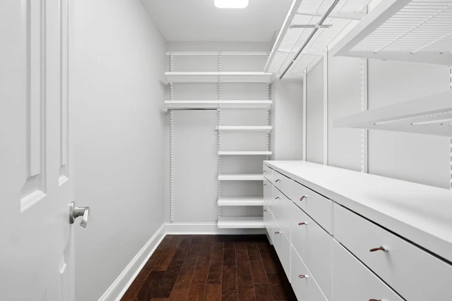 Narrow white walk-in closet with tall shelving on the left and drawers along the right, dark wood floor visible at the bottom.