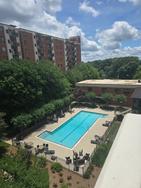 Apartment building with balconies overlooks a fenced swimming pool and landscaped courtyard under a cloudy sky.
