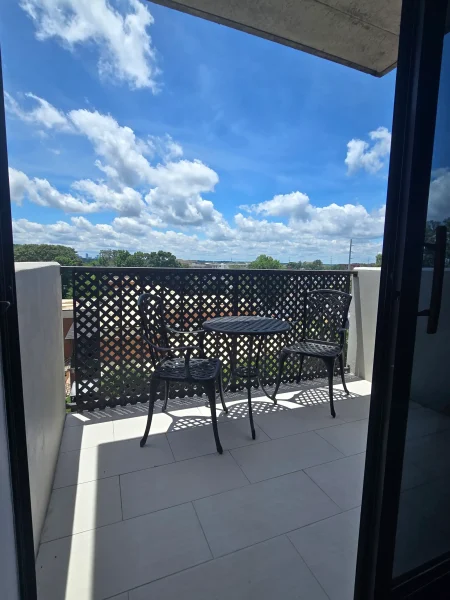 Outdoor balcony with a black metal lattice railing, round table and chairs on light tile, bright blue sky with white clouds above.