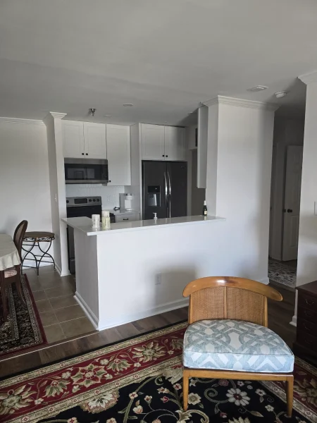 Open kitchen with white cabinets, stainless steel fridge, and a white island; a patterned rug and a wicker chair nearby.