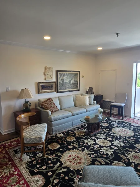 Living room with a light blue sofa, decorative pillows, a glass coffee table, and a dark floral area rug; wall art and a lamp on a side table nearby.