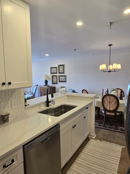 White kitchen with marble countertop island, stainless-steel sink, and built-in dishwasher; dining area visible in background with chandelier and framed art.