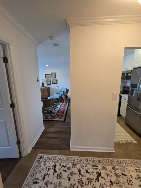 Hallway opening into a living room with framed wall art, wooden furniture, and a red-patterned rug viewable at the end.