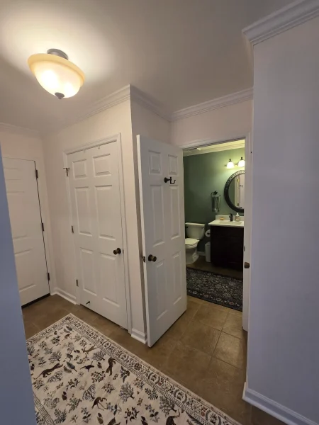 Hallway with white paneled doors; open doorway reveals a green-walled bathroom with vanity, mirror, and toilet, under a ceiling light.