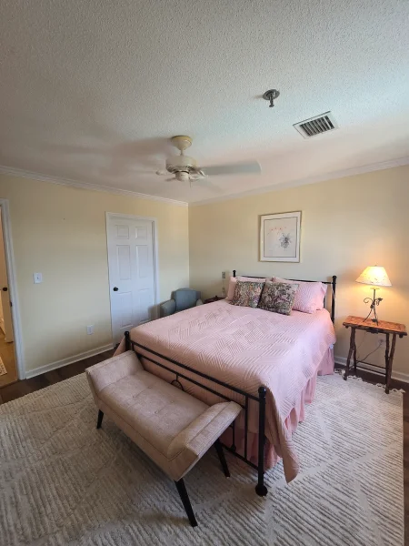 Bedroom with pale yellow walls, pink quilted bed, decorative pillows, and a bench at the foot of the bed.