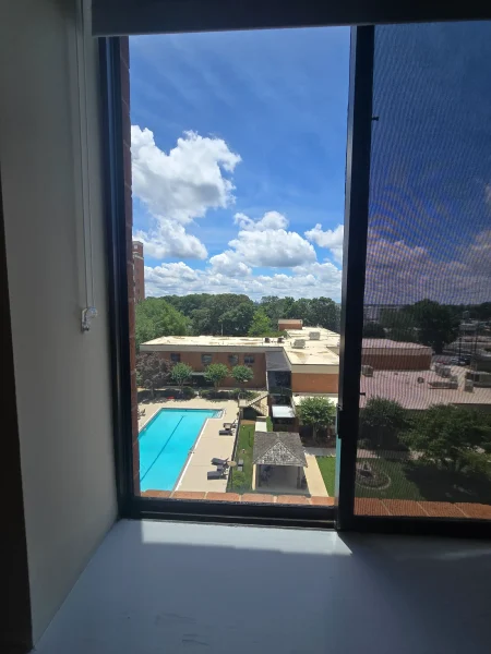 View from an open window: blue sky with white clouds over a rectangular swimming pool and surrounding brick buildings.
