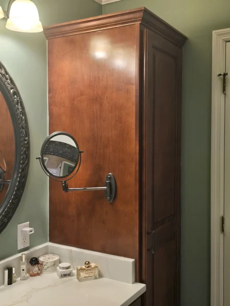 Wooden bathroom cabinet above a marble countertop with a round magnifying mirror mounted on an adjustable arm; vanity items scattered on the counter.