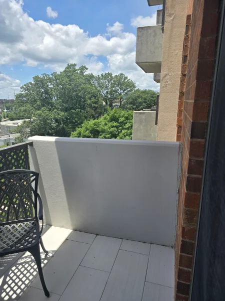 Balcony with light-colored tiled floor, a black lattice chair, white railing, and a brick wall along a concrete building, overlooking green trees and a blue sky with clouds.