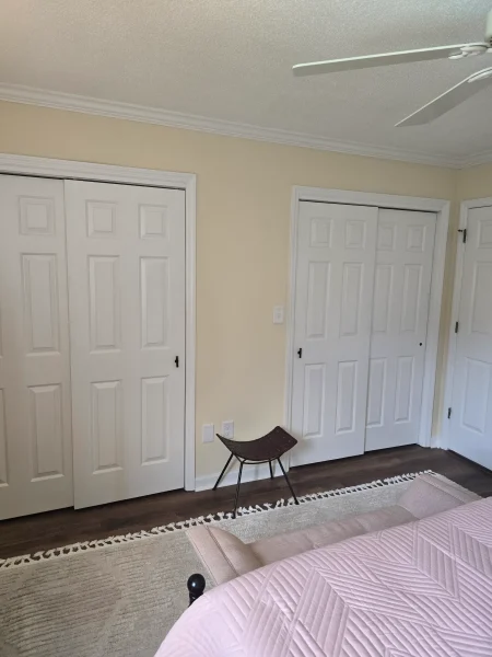 Bedroom with beige walls, white panel doors, and a ceiling fan; a small woven chair near the wall, rug along the floor, and a pink quilted bed in foreground