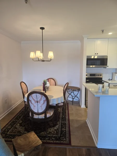 Round dining table with four upholstered chairs on a patterned rug; a chandelier hangs above in an open kitchen area.