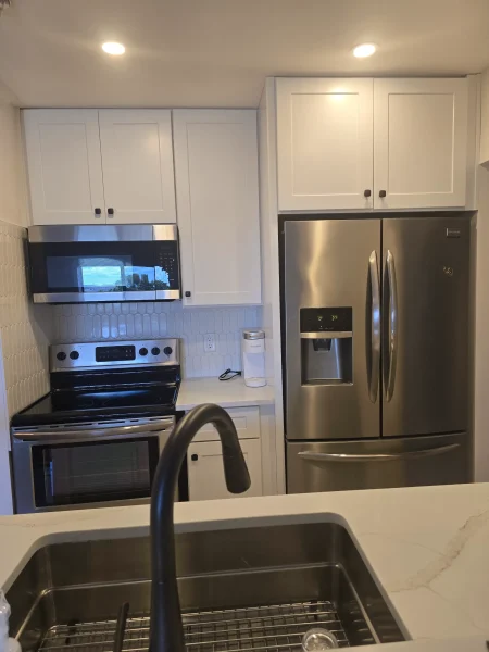 Stainless steel side-by-side refrigerator beside white upper cabinets in a bright kitchen with a black faucet in the foreground.