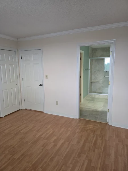 Empty room with light wood floors, beige walls, and white paneled doors on the left; doorway on the right leads to a tiled bathroom with a glass shower enclosure.