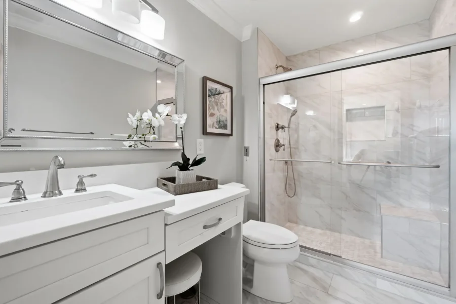 Modern bathroom with a white double vanity, silver faucets, an orchid in a tray, and a marble-tiled glass shower.