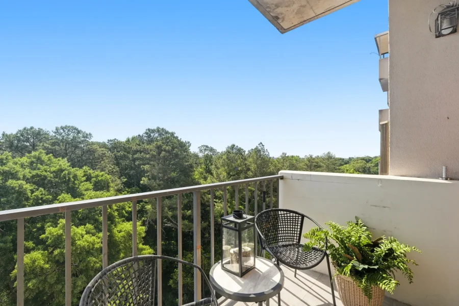 Balcony seating area with two black wicker chairs and a glass-topped table, lantern, and potted plant, overlooking trees under a clear blue sky.