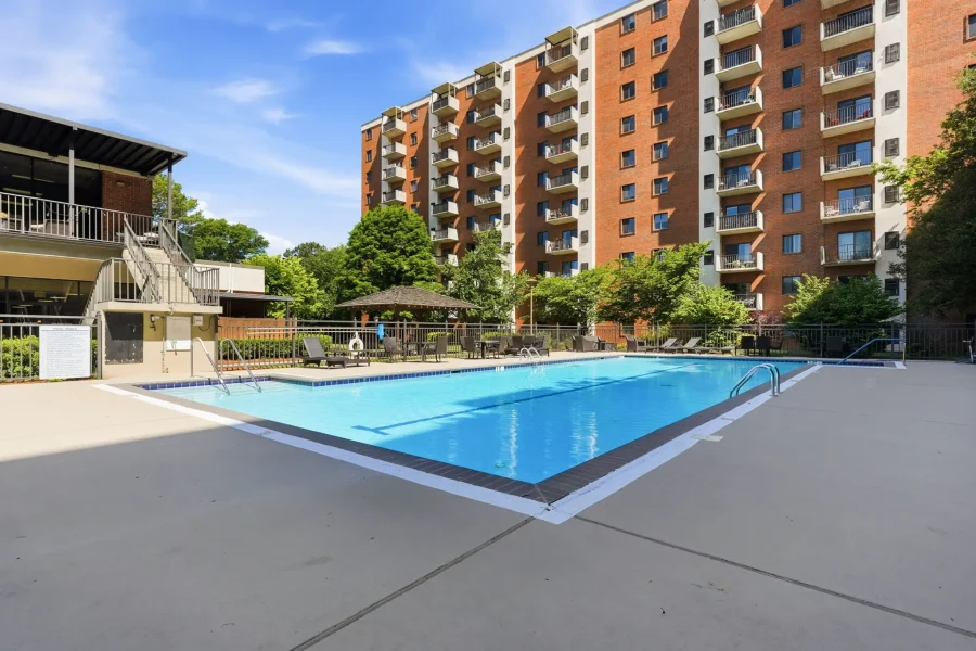Poolside at a residential complex: a rectangular blue pool with lounge chairs and a gazebo, set against a tall red-brick apartment building with balconies under a blue sky.