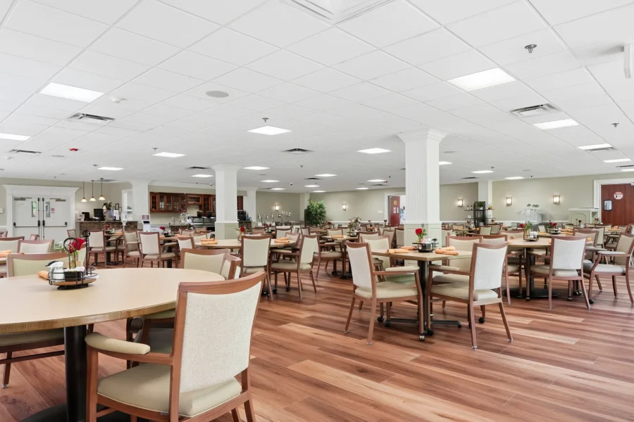 Bright cafeteria dining area with many round and rectangular tables, beige chairs, and wood flooring under a white tiled ceiling with columns.