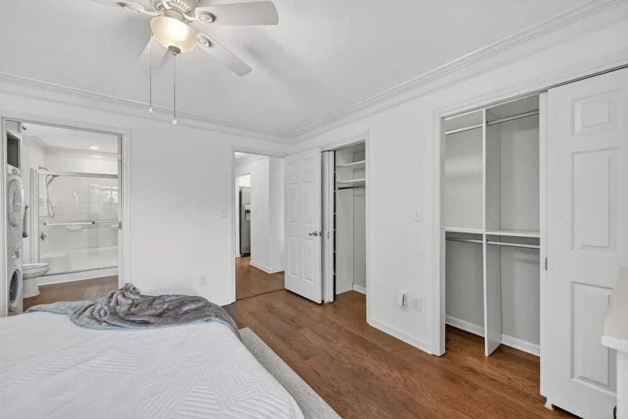 Bright bedroom with white walls and a ceiling fan, open closets, and a visible bathroom with a glass shower; laundry appliances nearby.