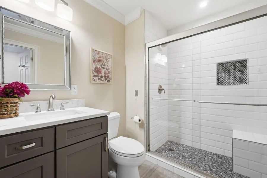 Dual-sink bathroom vanity with a framed mirror, pink flowers, and a glass shower enclosure with white subway tiles and a mosaic floor outside the shower stall edge.