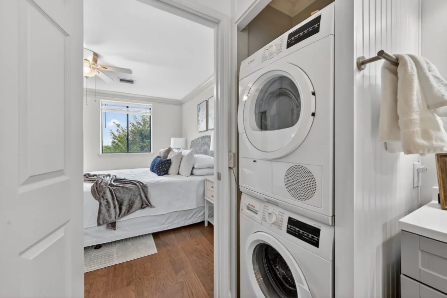 Stacked washer and dryer in a hallway closet, opening to a bright bedroom with a made bed and window.