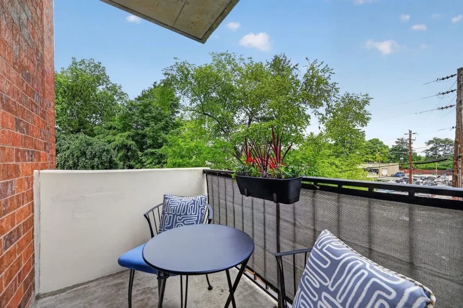 Relaxing balcony scene with a round blue table, metal chairs, patterned cushions, and a planter of red-leafed plants on a railing against a backdrop of trees and blue sky