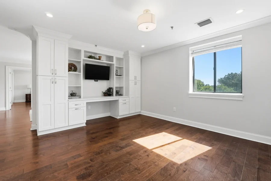 White built-in entertainment wall with shelves, cabinets, and a wall-mounted TV in a bright living room with dark hardwood floors.