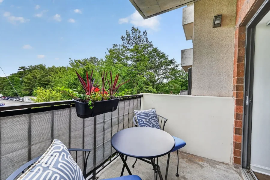 Balcony seating area with a round metal table and blue cushions, a planter with red-tipped plants, and a view of trees and parked cars beyond the railing.