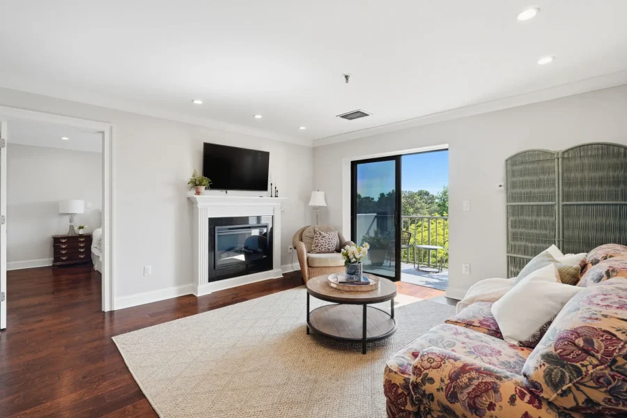 Bright living room with a white fireplace and wall-mounted TV, wicker chair, round coffee table, and floral sofa; sliding glass door to a balcony with greenery.