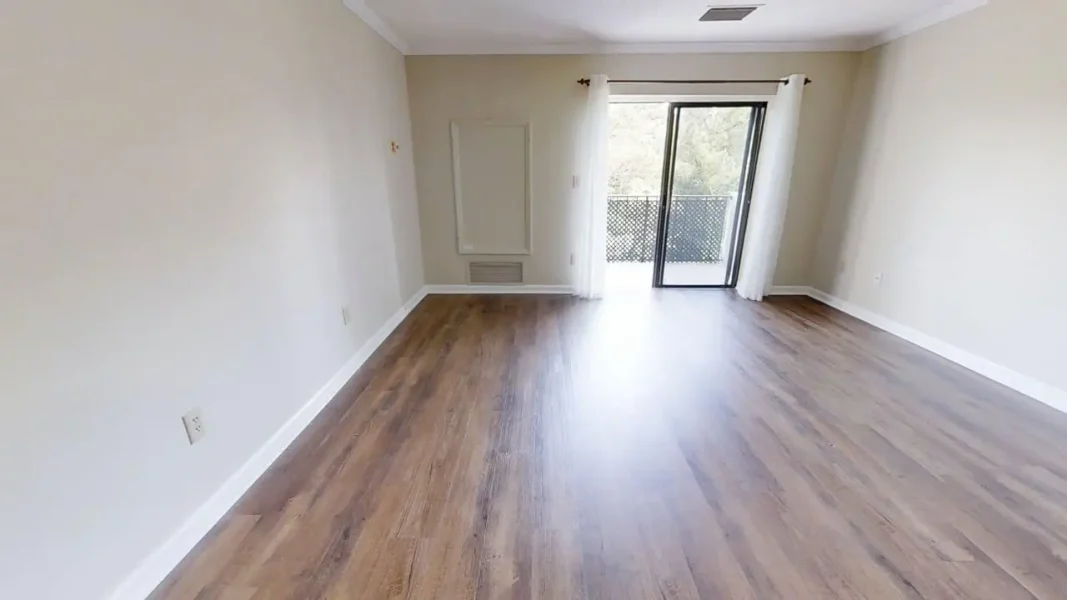 Empty living room with light wood flooring, beige walls, and white baseboards; sliding glass door with sheer white curtains opens to a balcony.