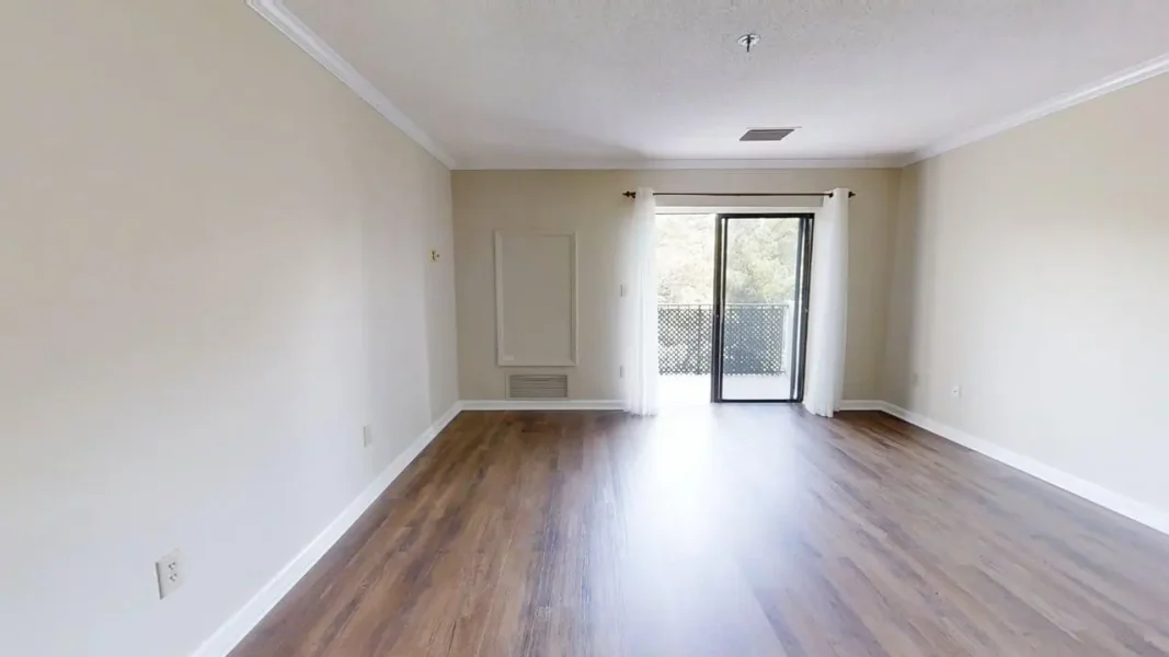 Empty living room with wood floors, beige walls, and a sliding glass door with sheer curtains to a balcony there.