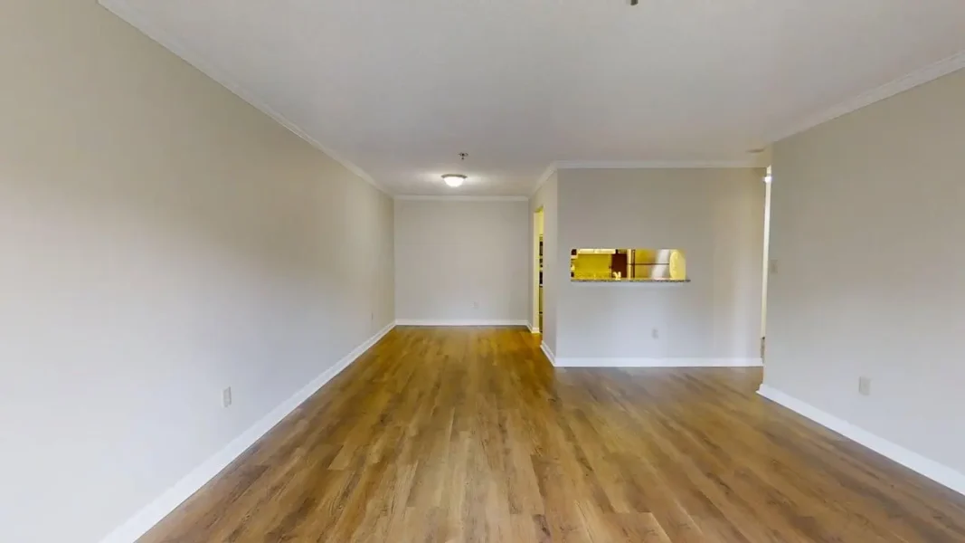 Empty living room with light beige walls, white baseboards, and wood-look flooring; open doorway to kitchen with pass-through window.