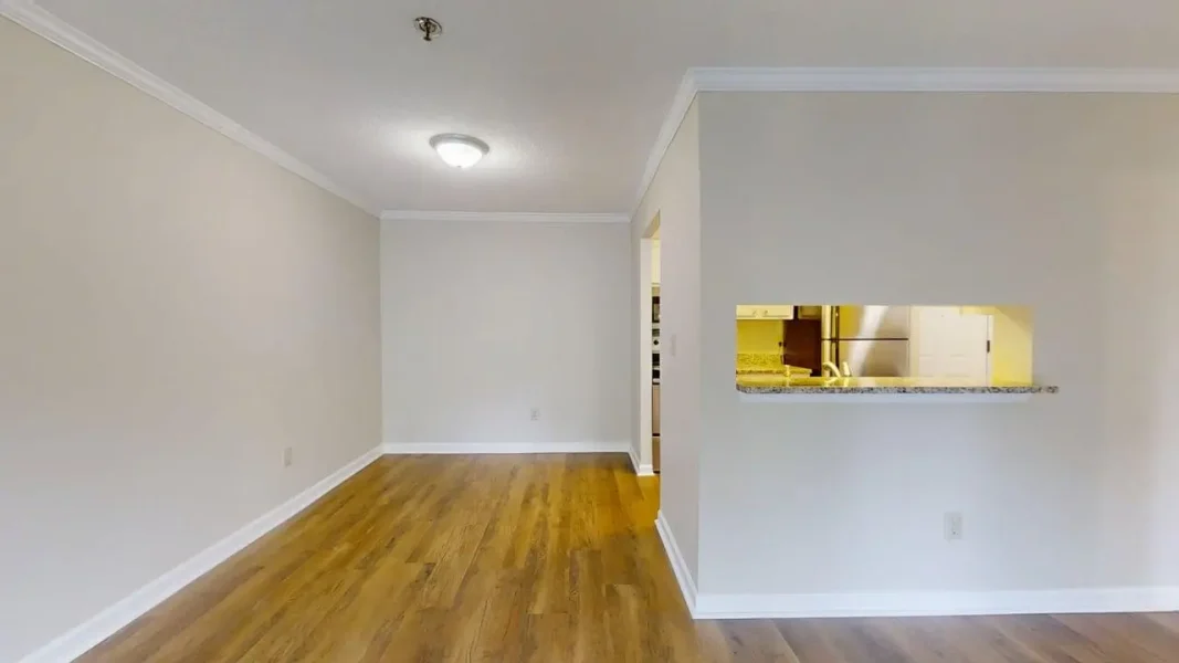 Empty living room with light beige walls, white baseboards, and hardwood flooring; a kitchen pass-through window on the right.