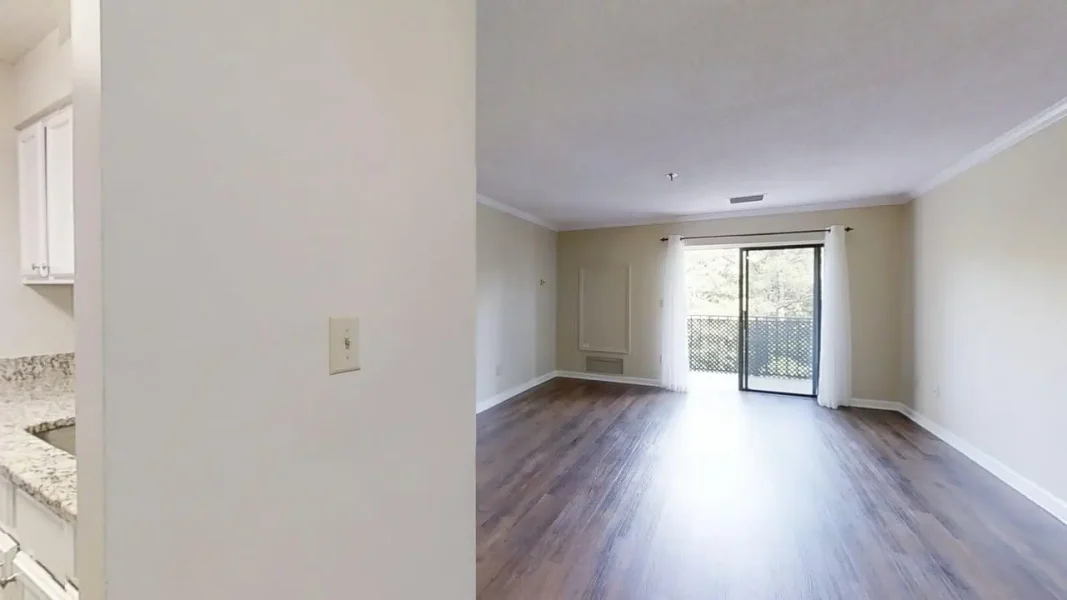 Bright, empty living room with wood-look flooring, light beige walls, and a sliding glass door to a balcony with white curtains on either side.