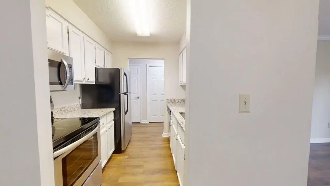 Galley-style kitchen with white cabinets, granite countertops, and stainless steel fridge and oven, viewed from a doorway.