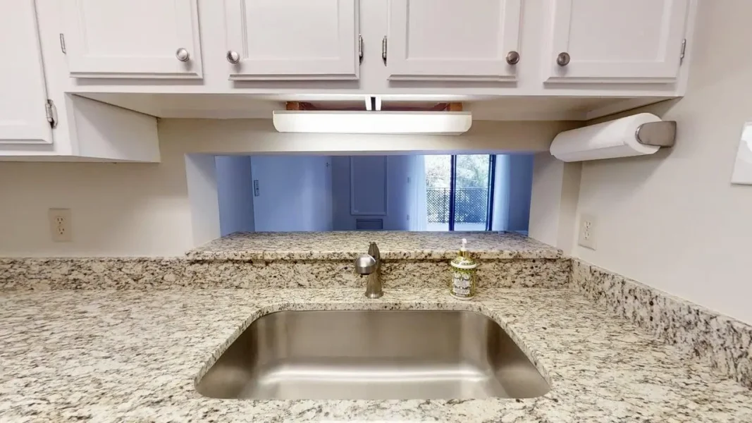Kitchen counter with granite surface, stainless steel sink, and soap dispenser; pass-through opening to the next room.