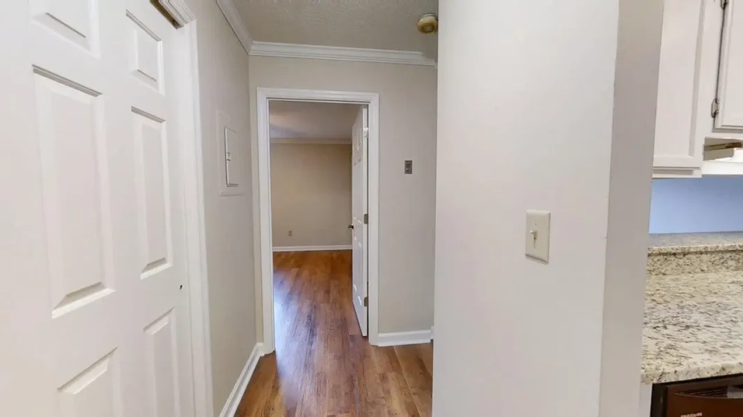 Hallway in a home with white doors, light gray walls, and wood flooring; open doorway at the end leads to another room, with a granite kitchen counter visible on the right.