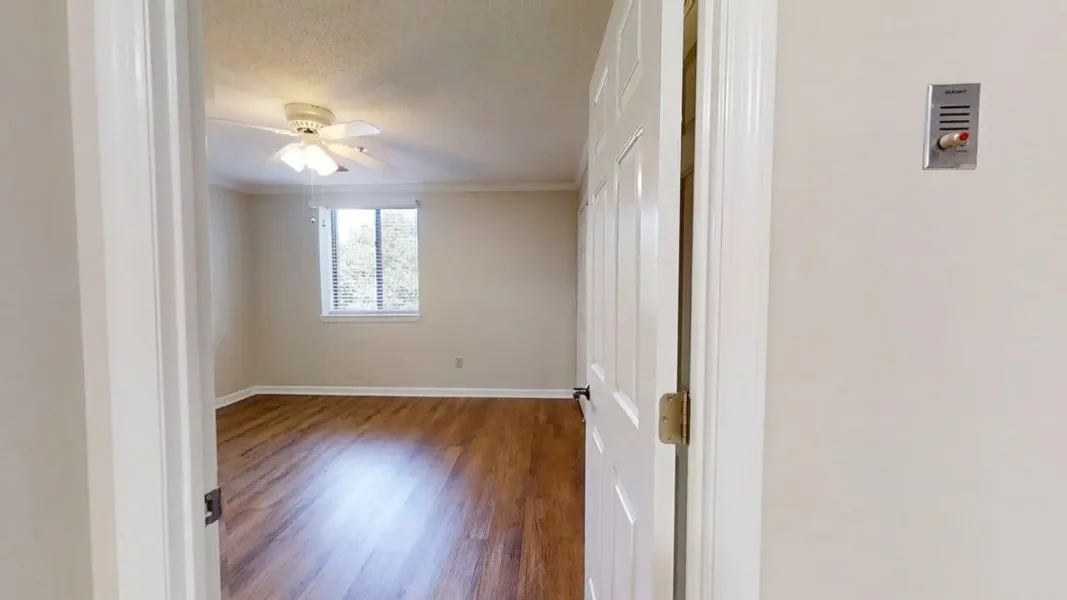 Empty living room with hardwood floors, beige walls, and a ceiling fan viewed from doorway; blinds-covered window at far wall.